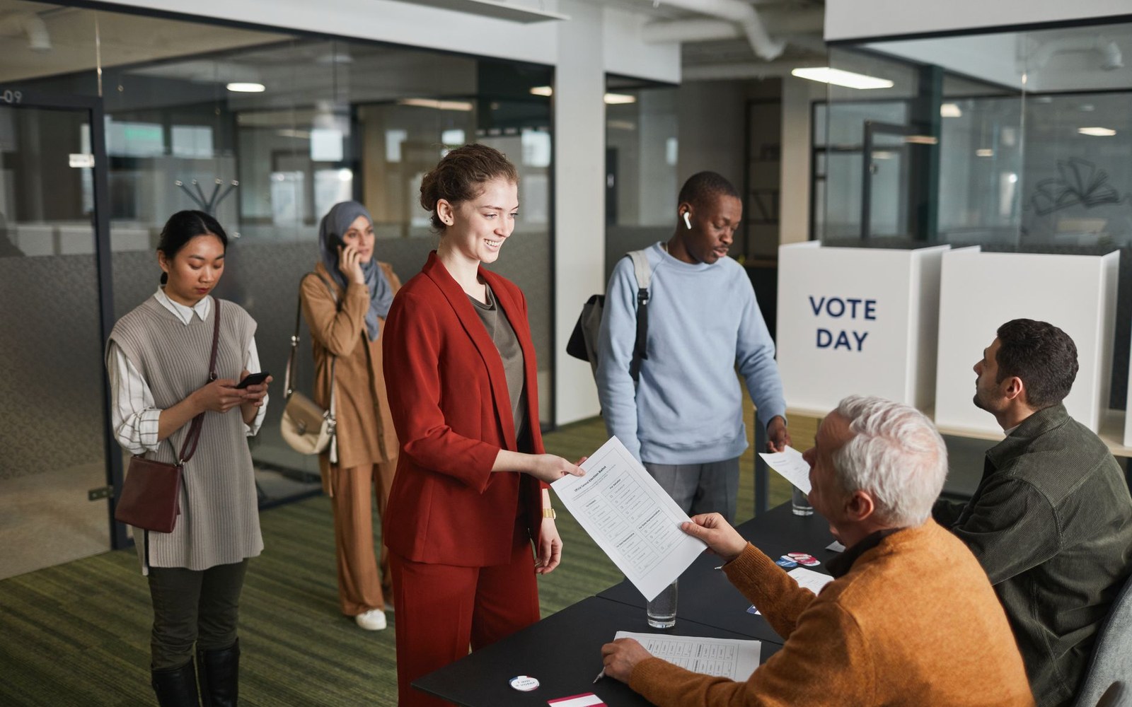People line up indoors to receive ballots at a table beside a "vote day" sign