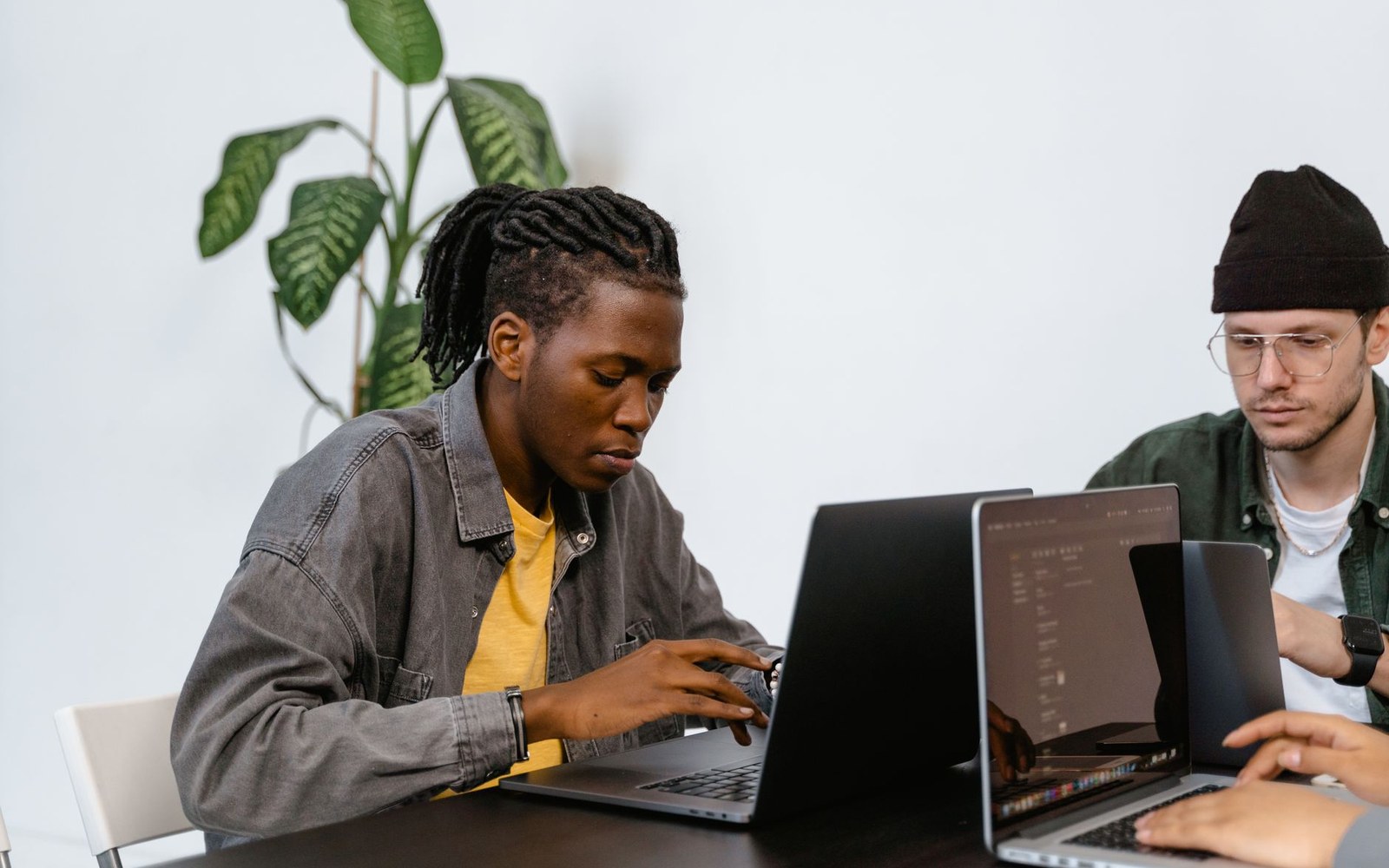 Two men sit side by side at a table, typing on separate laptops in an office