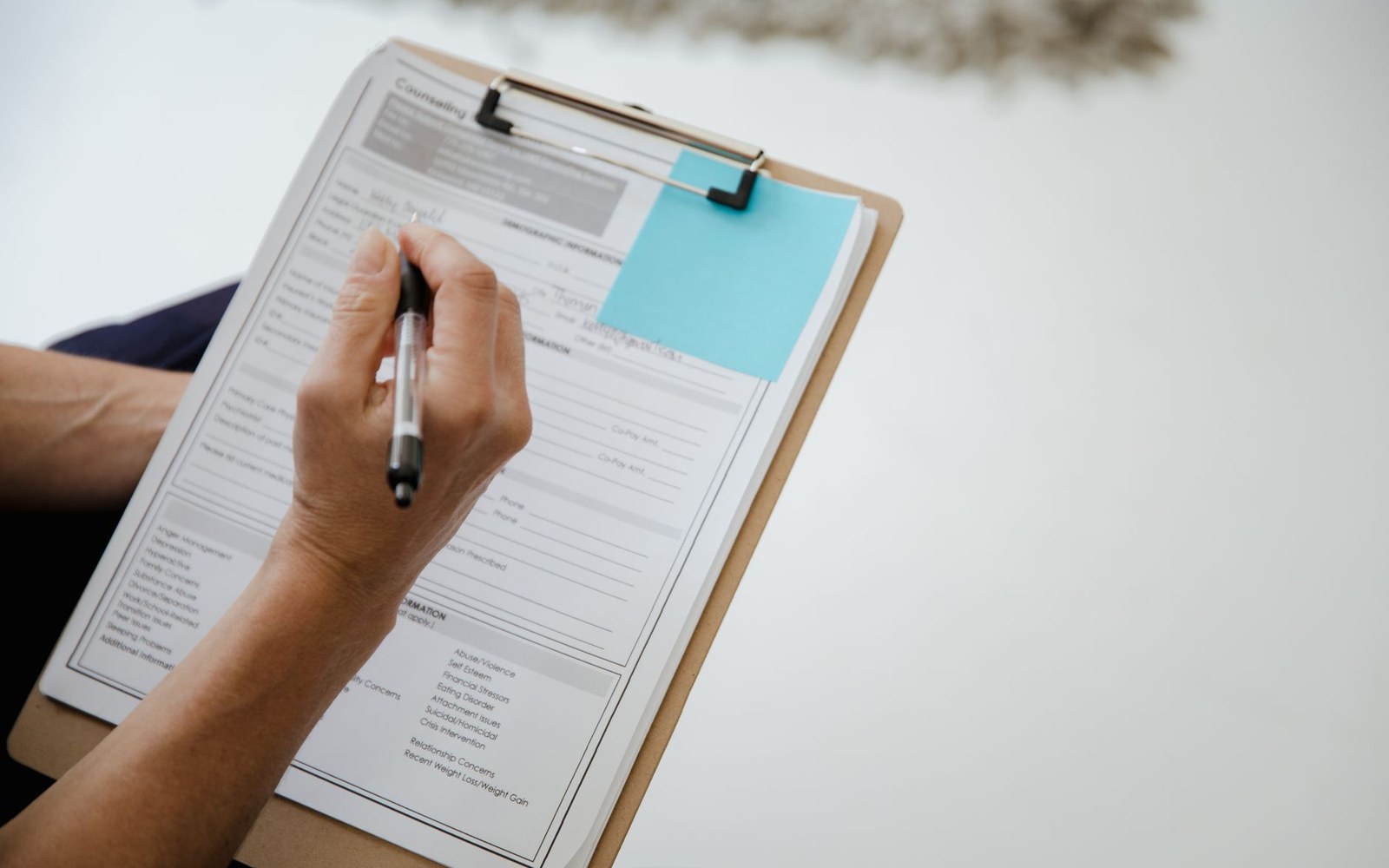 A hand fills out a counseling intake form on a clipboard with a sticky note attached