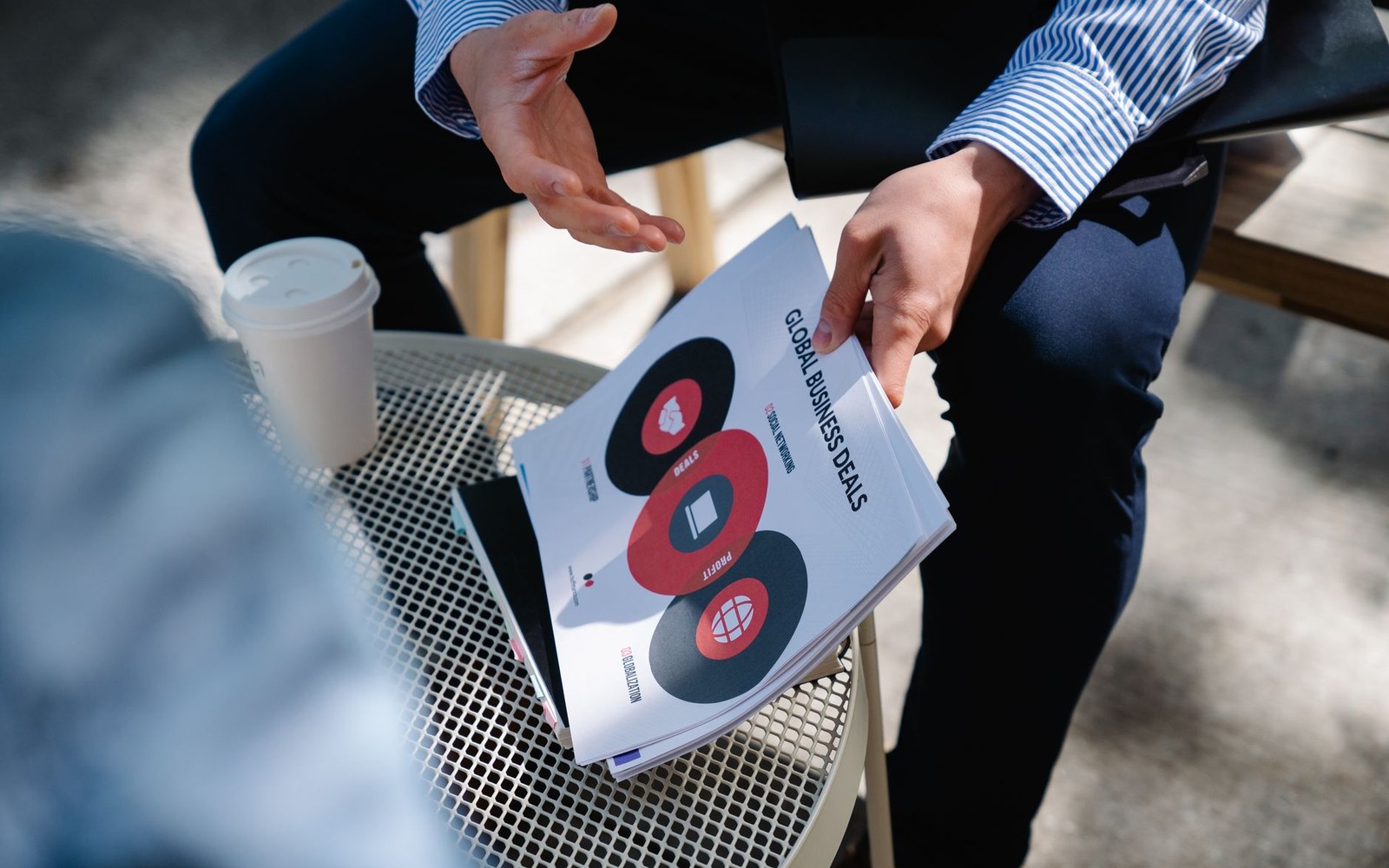 A person holds a "Global Business Deals" report while seated at an outdoor table with coffee