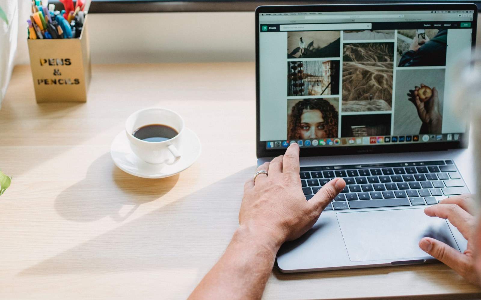 Hands typing on a laptop showing a photo gallery, beside a coffee cup and pen holder